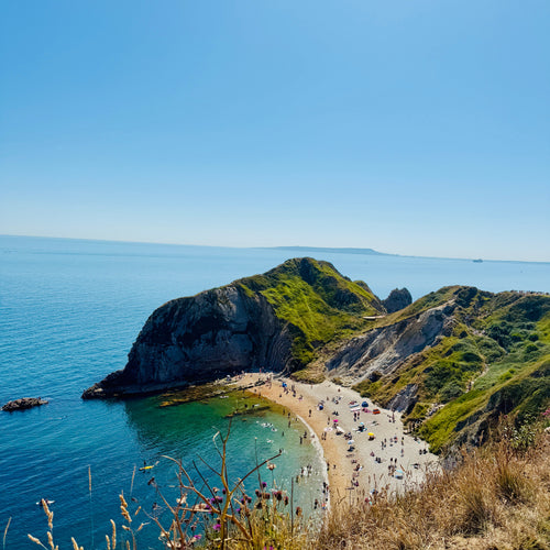 Beach with people on a clear day, surrounded by green hills and blue water.